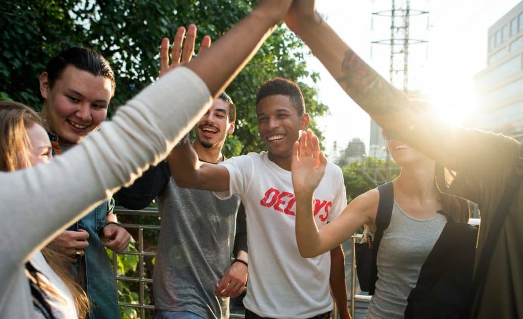 A group of friends are giving each other high fives to cheer each other on and having fun together. They are standing on a bridge outside in front of some trees.