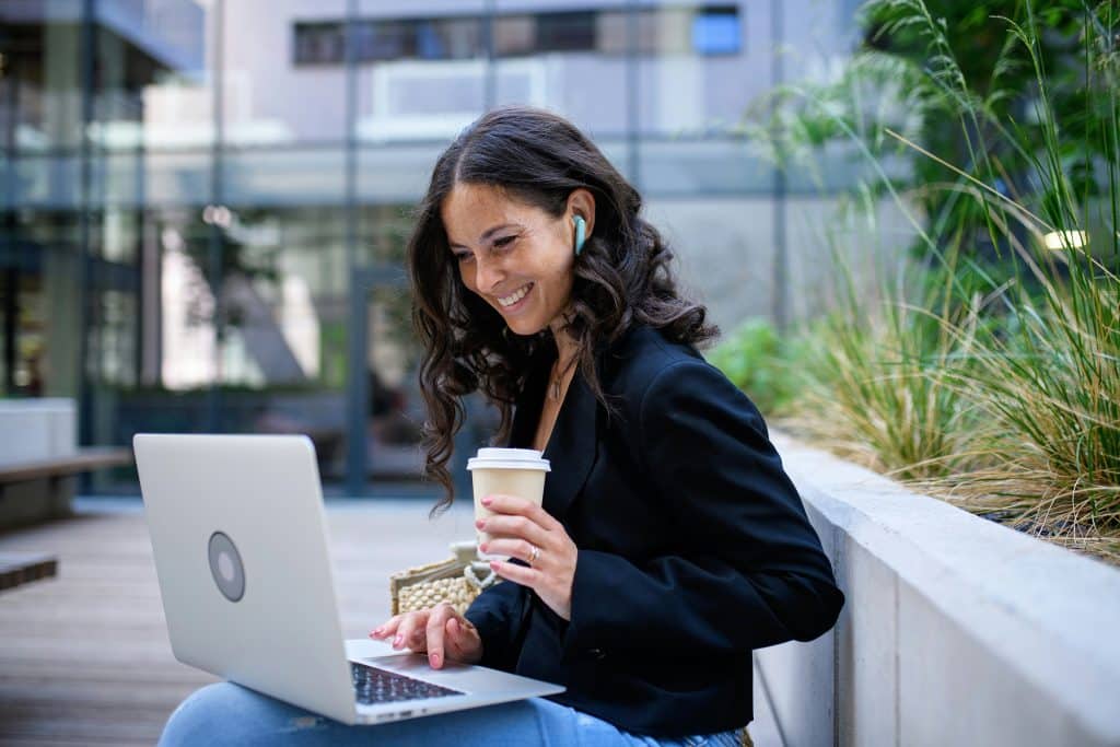 A stylish young businesswoman is working outdoors on her laptop, on the forecourt of her office building. She is holding a coffee in her hand and wearing headphones. She is wearing a black jacket and jeans.