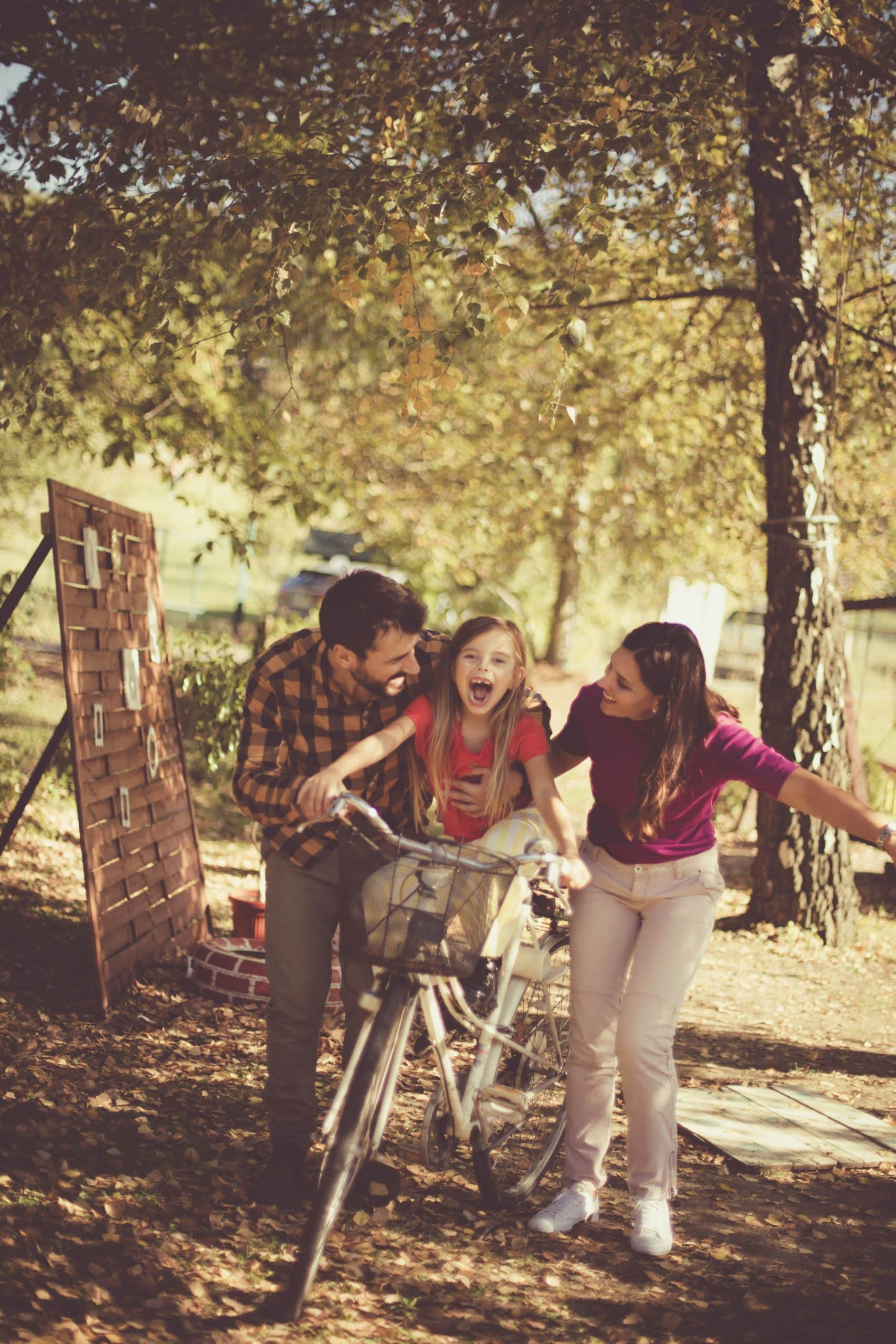 A couple teaches their daughter to ride a bicycle, standing on either side of her. They are outside in a park or garden, under the trees.