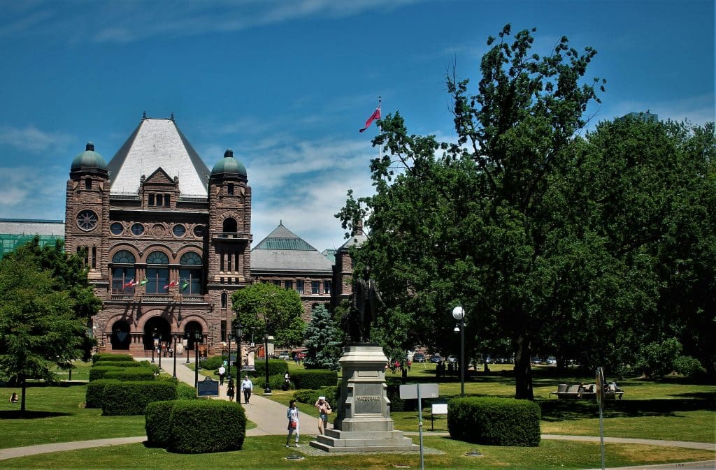 University of Toronto in Canada on a fine day. The building is constructed of brown brick and a large garden with trees surrounds the school.