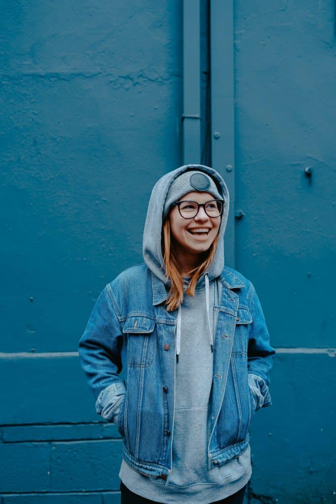 A young blonde woman wearing glasses is dressed entirely in blue denim and poses in front of a wall that is also entirely blue.